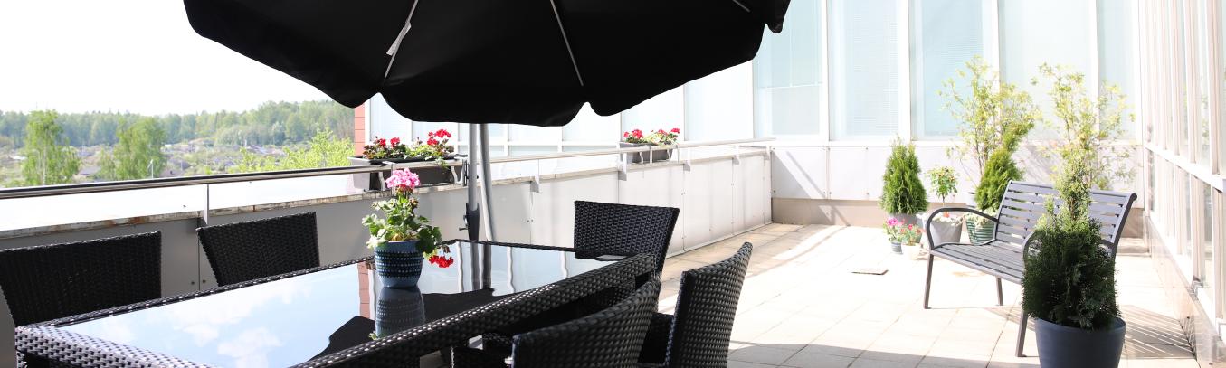 Aurinkoinen terassi, jolla mustat terassikalusteet ja iso aurinkovarjo sekä paljon kukkia ja viherkasveja. Taustalla näkyy puistomaisemaa.